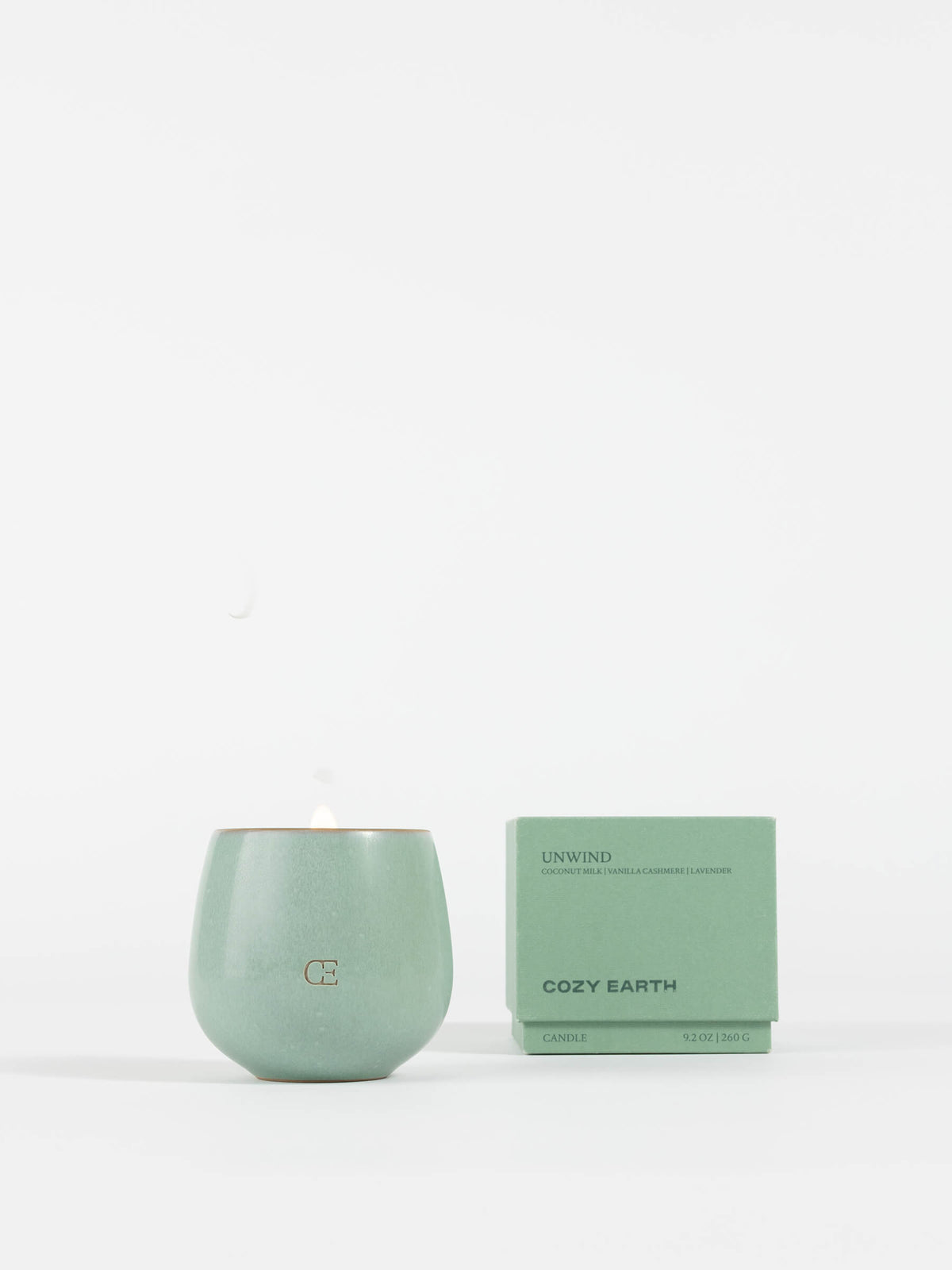 A pale green Cozy Earth Candle UNWIND in a round container, with a small flame, sits beside its matching box labeled "UNWIND" and "Cozy Earth" on a plain white background.
