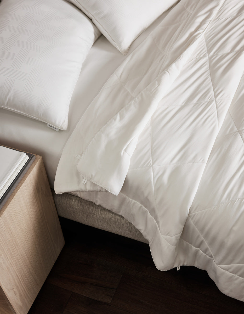 A neatly made bed features the Cozy Earth Bamboo Insert Bundle—white quilted comforter and pillows—partially pulled back. A light wooden nightstand with a closed book sits beside the bed on a dark wood floor.|Filling:Extra