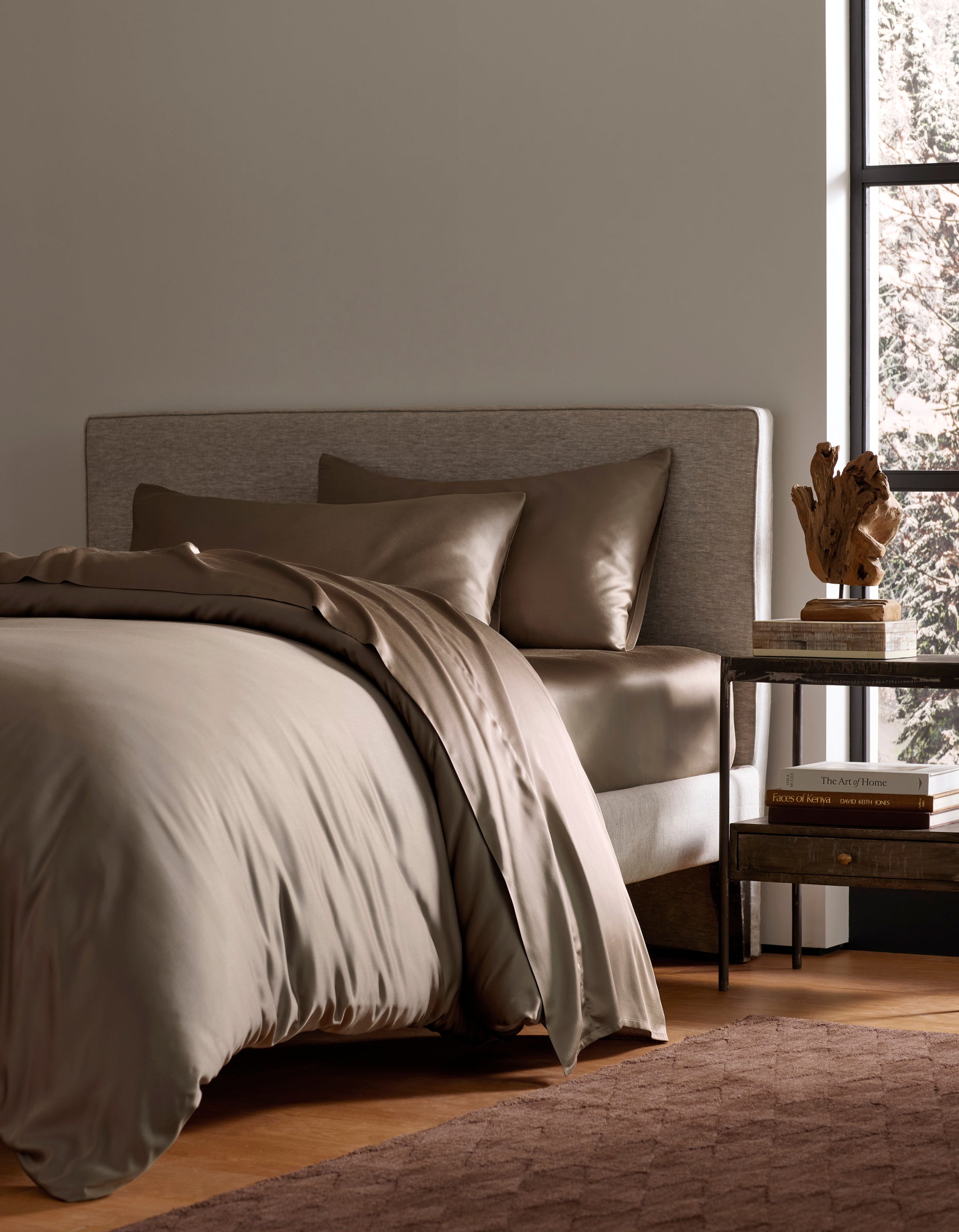A modern bedroom features a neatly made bed with Cozy Earth’s Bamboo Bedding Deluxe Bundle in taupe, paired with pillows. A wooden nightstand nearby holds books, a lamp, and decor, as sunlight adds warmth to the inviting space.|Color:Walnut