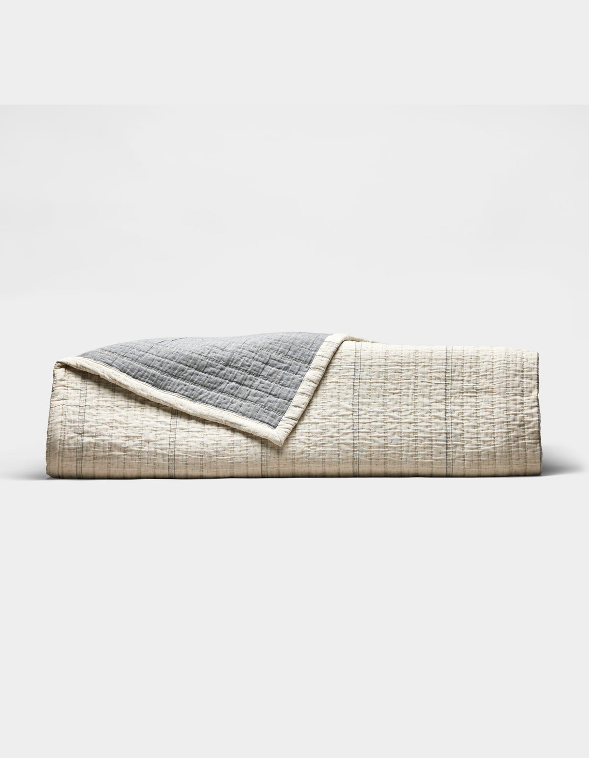 The Cozy Earth Baja Matelasse Coverlet features a neatly folded design with a cream side adorned with subtle lines and a textured gray reverse, shown against a plain white background.|Color:Alabaster/Oceanside