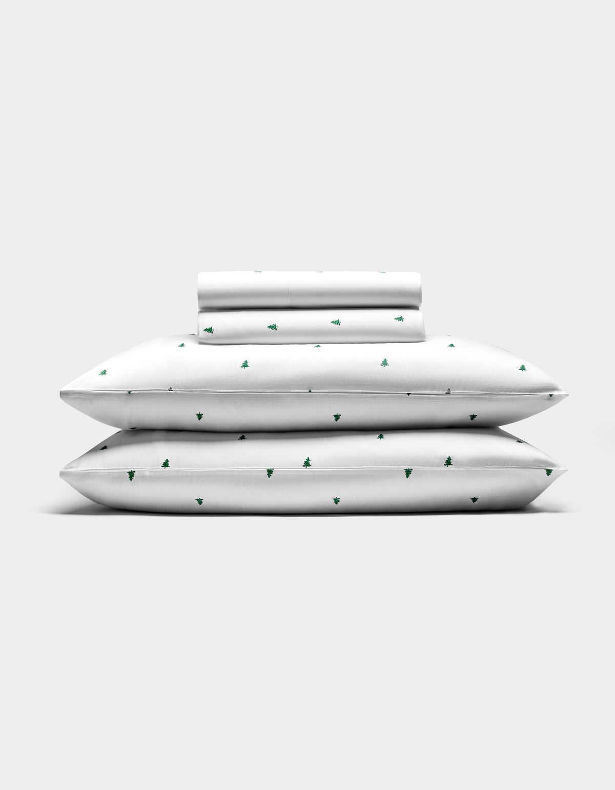 The Cozy Earth Bamboo Sheet Set features two pillows, a folded sheet, and a rolled pillowcase—all white with small green tree patterns—arranged against a plain light gray background.|Color:Jade Tree Snow