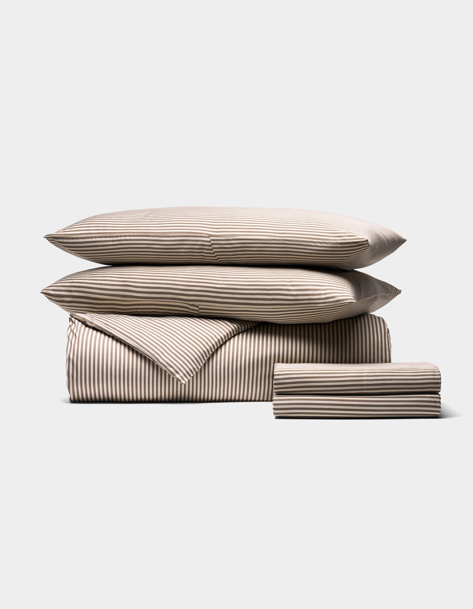 The Bamboo Bedding Deluxe Bundle by Cozy Earth features a neatly stacked beige and white striped set with two pillows, a folded comforter, and a folded sheet arranged on a plain light background.|Color:Classic Stripe in Ebony