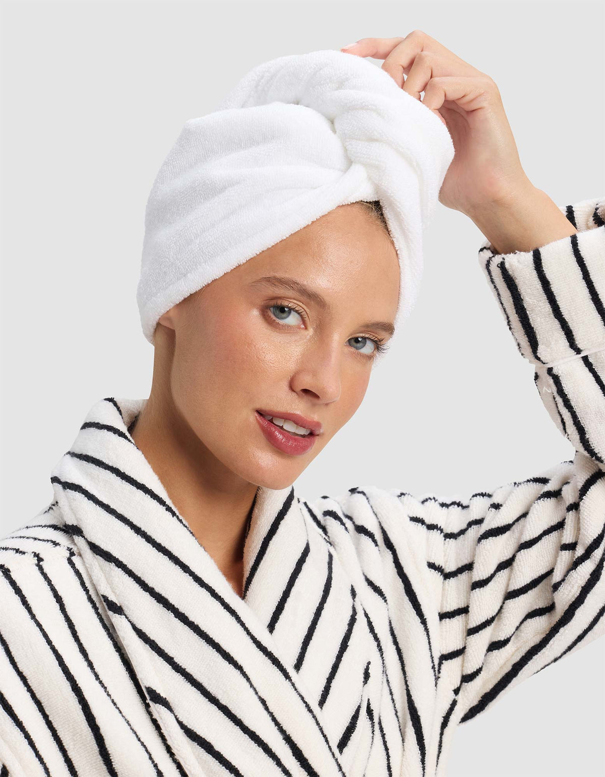 A woman smiles softly, touching the Cozy Earth Luxe Hair Towel wrapped around her head, while wearing a white bathrobe with black stripes against a plain light background. |Color:White