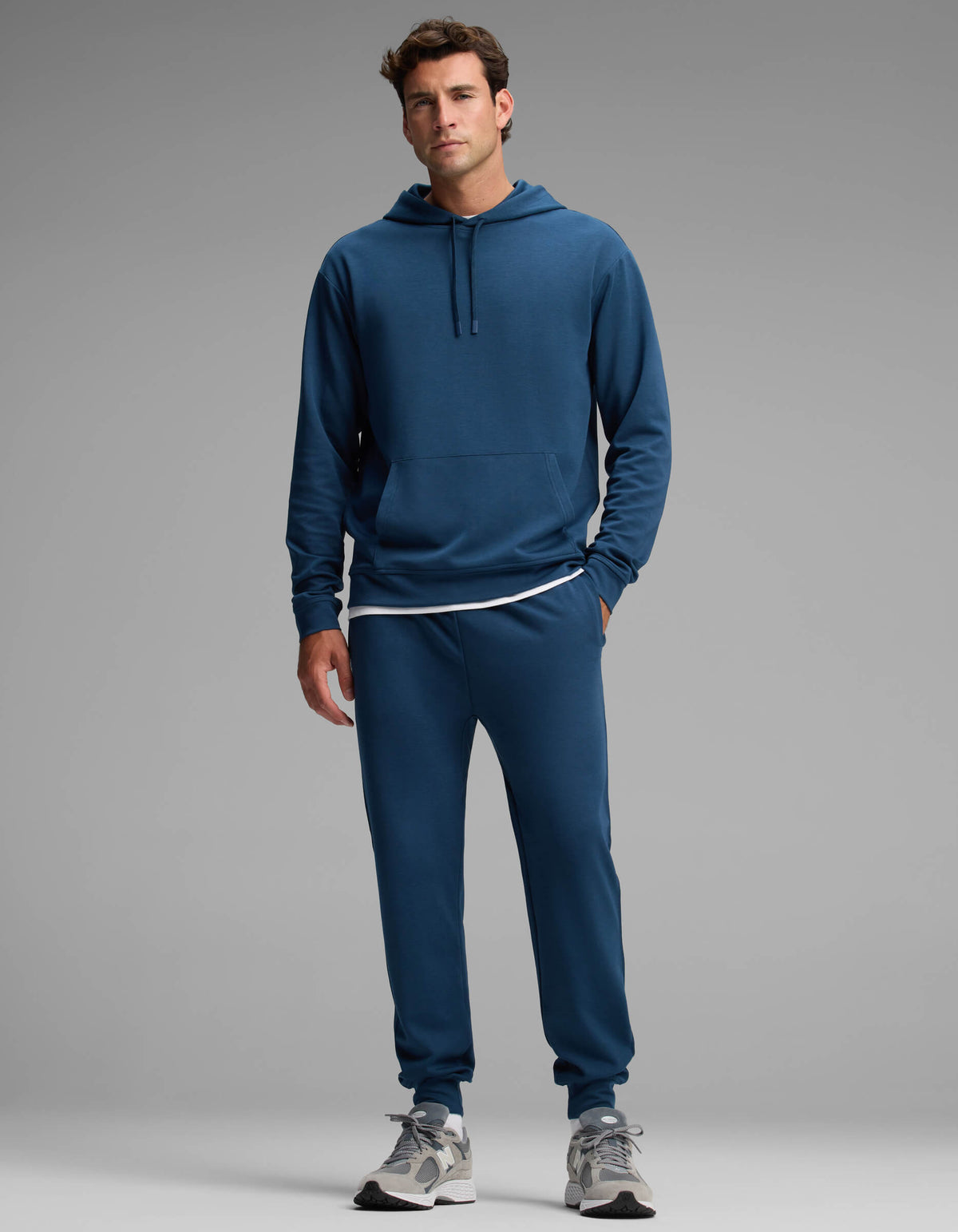 A man stands against a gray background wearing the Cozy Earth Men's Versa Hoodie & Jogger Set in blue, paired with a white T-shirt and gray sneakers. He looks forward with a neutral expression, one hand by his side and the other slightly bent.|Color:Obsidian