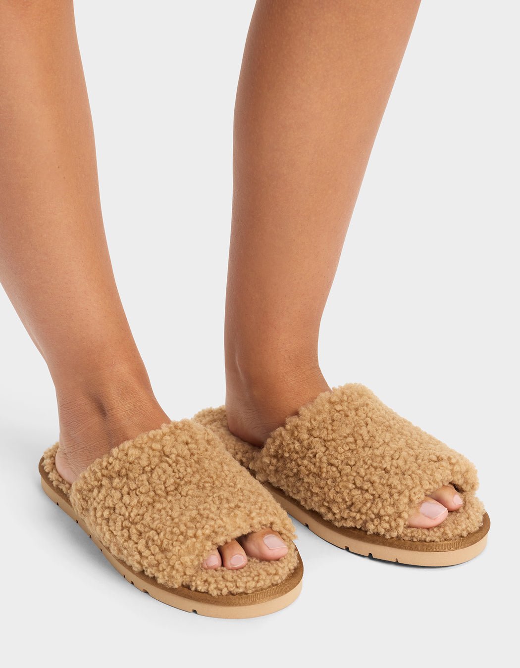 A person wears Cozy Earth’s Puffy Sheep Slides—fuzzy, tan open-toe slippers with thick, textured uppers and flat soles—standing on a white background.|Color:Fawn