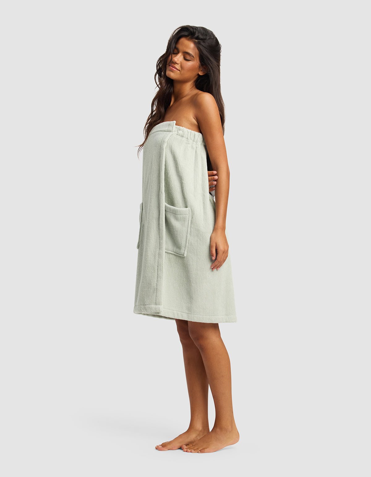 A woman with long dark hair stands barefoot, modeling the Cozy Earth Ribbed Terry Bath Wrap—light-colored, strapless, with two large front pockets—against a plain light gray background.|Color:Laurel