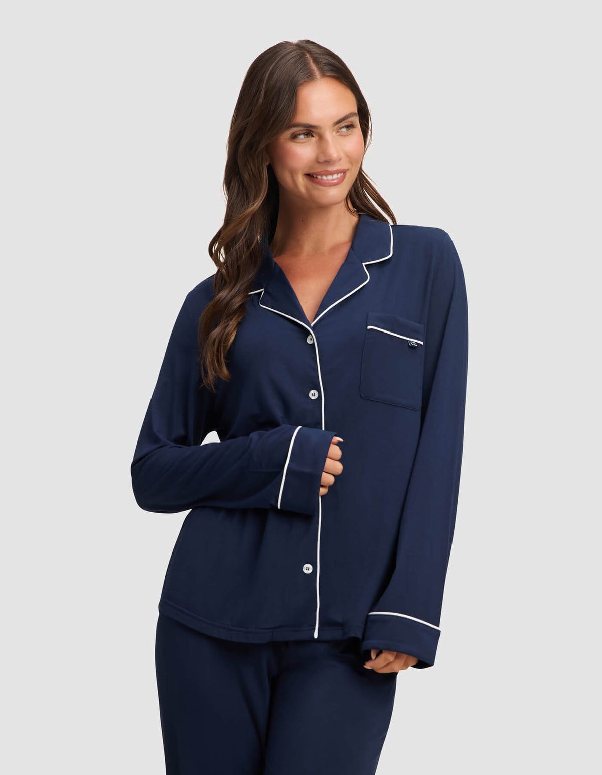 A woman with long brown hair smiles in a Cozy Earth Women's Bamboo Stretch-Knit Long Sleeve Pajama Top, navy blue with white piping, paired with matching pants, against a plain light gray background. |Color:Navy