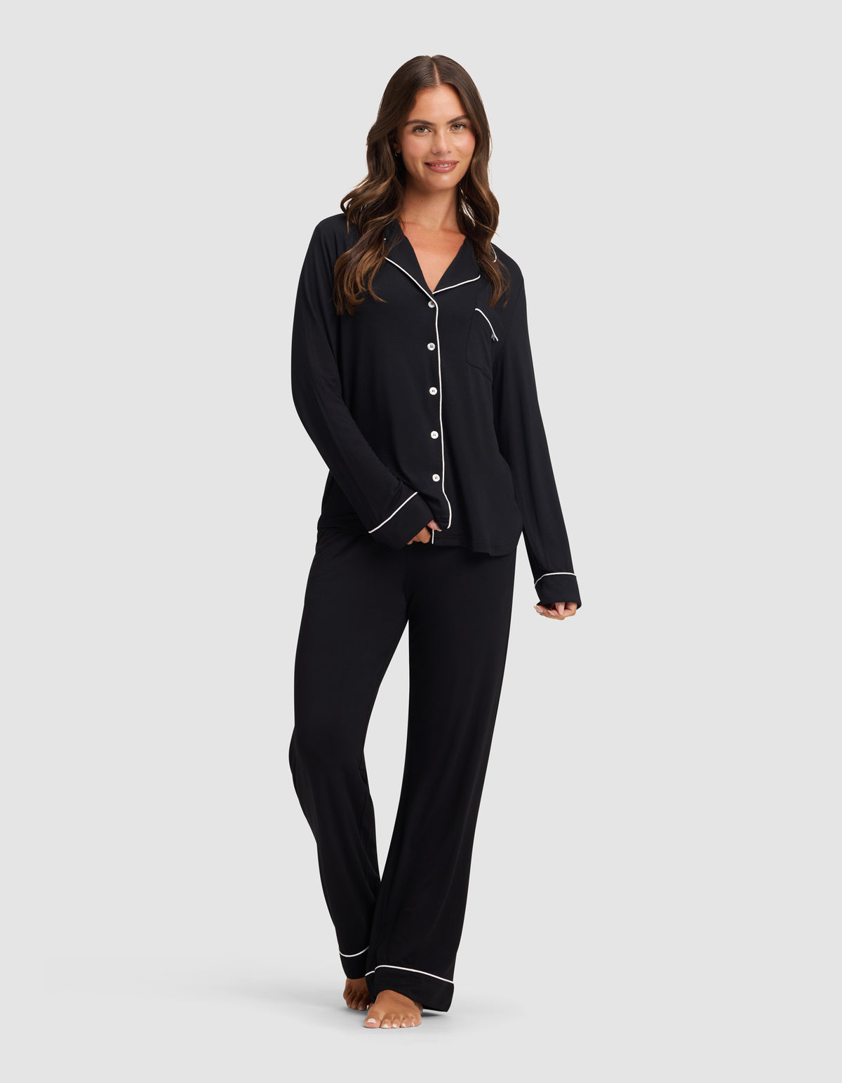 A woman with long brown hair, barefoot and smiling, models the Cozy Earth Women's Bamboo Stretch-Knit Pajama Pant in black with white piping, paired with a matching button-up top, against a plain light gray background.|Color:Black