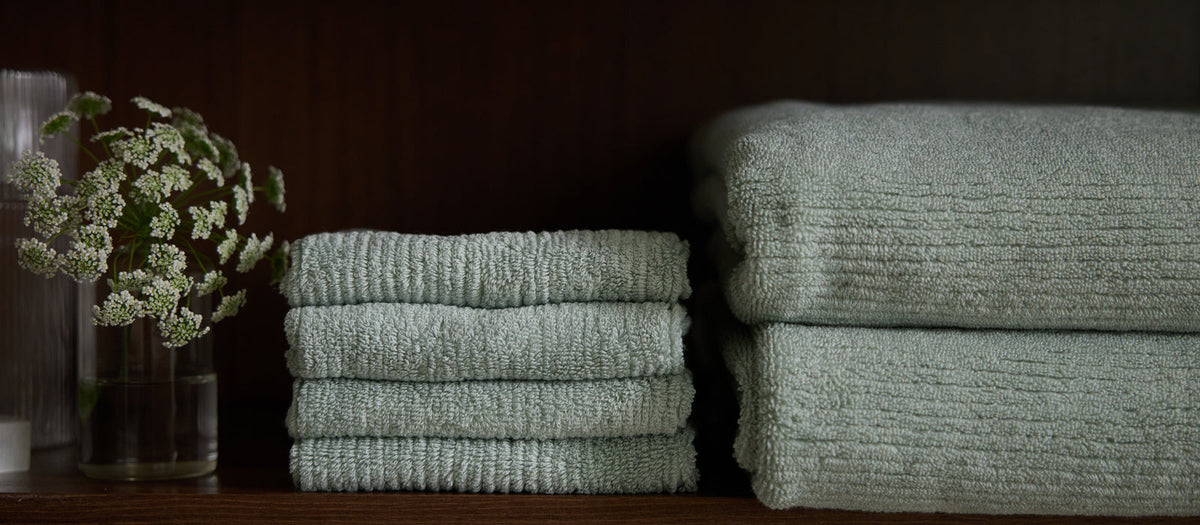 A stack of green Ribbed Terry Washcloths and 2 Ribbed Terry Towels next to a small vase of flowers