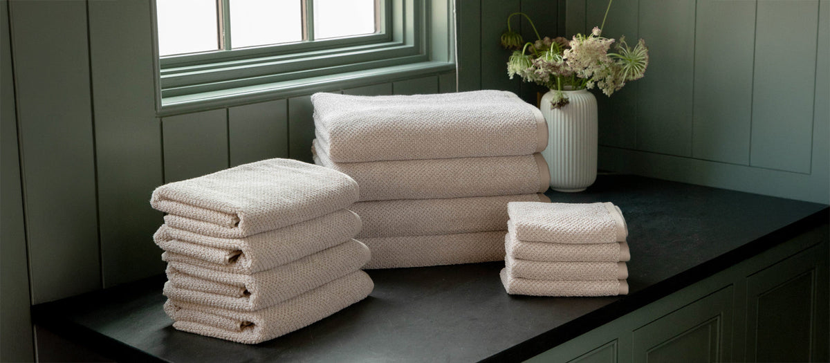 A stack of Cozy Earth Nantucket Bath Towels, Hand Towels, and Washcloths sit on a countertop under a window. A vase with flowers and green paneled walls are also shown.