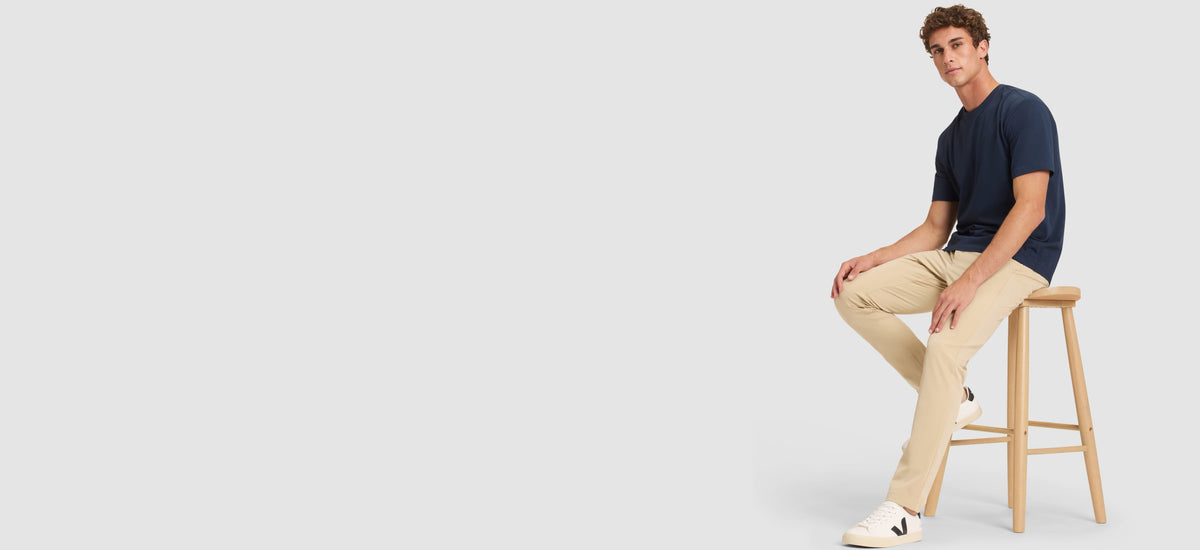 Man sitting on a stool wearing beige pants and a navy shirt against a white background