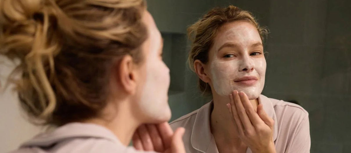A woman applies a mask to her face in a mirror