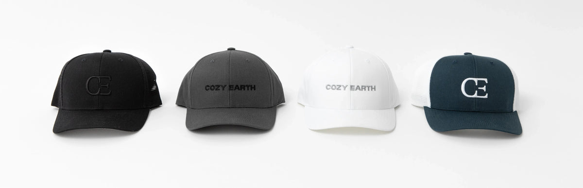 Four baseball caps are displayed in a row on a white background. The caps are in black, gray, white, and dark green colors, respectively. The first and last caps have the "CE" logo, while the middle two caps have the text "Cozy Earth.