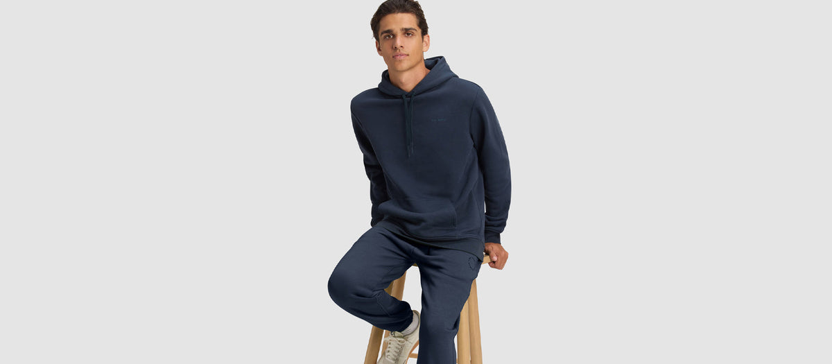 A man wearing Cozy Earth Cityscape 2.0 Hoodie & Sweatpant set sits on a stool in front of a light grey background.