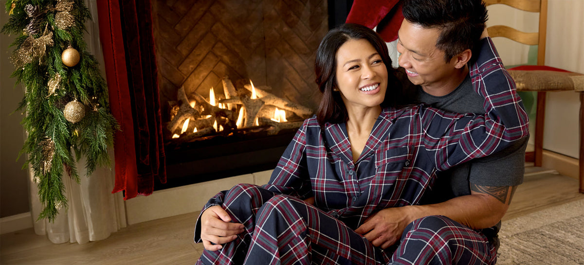 A man and woman in matching pajamas sit in front of a fireplace decorated with pine boughs