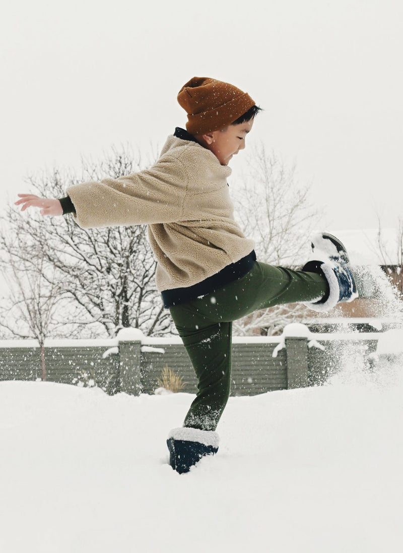 A young child wearing Cozy Earth's Children's Bamboo Waffle Knit Base Layer Pant plays outside, kicking up snow on a wintry day with trees and a fence in the background.