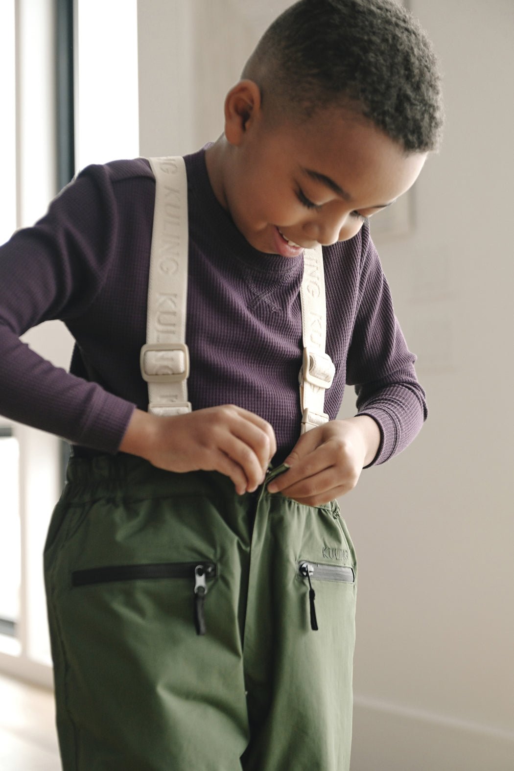 A young boy smiles while wearing Cozy Earth's Children's Bamboo Waffle Knit Base Layer Long Sleeve in purple and green pants with white suspenders as he zips up his pants.