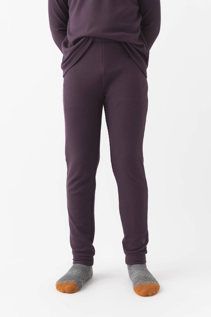 A child stands against a white background wearing Cozy Earth’s Children’s Bamboo Waffle Knit Base Layer Pant in Plum, paired with a matching top and gray socks with orange toes. Their hands are in their pockets, upper body partially out of frame.