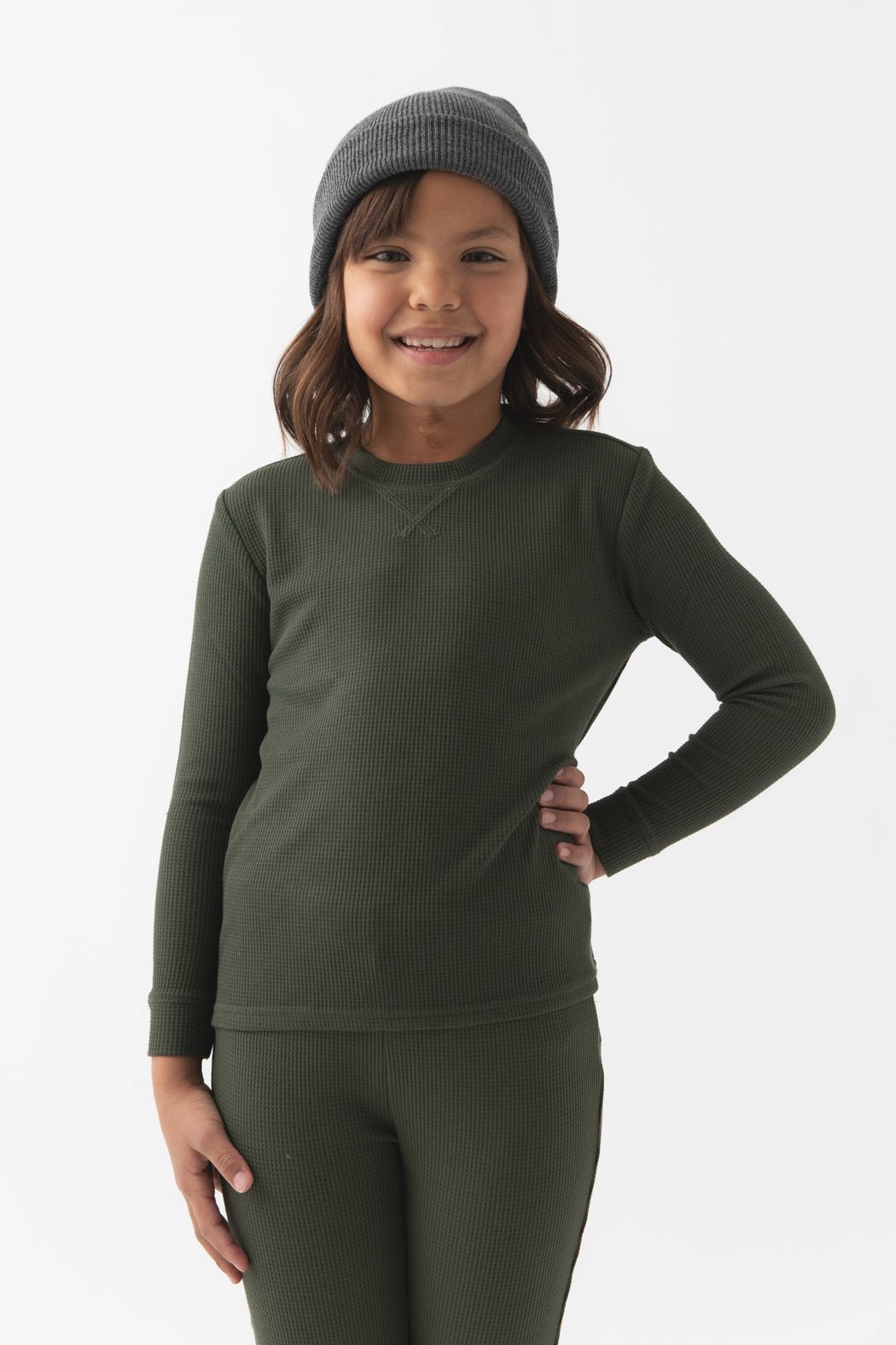 Smiling child with medium-length brown hair, wearing a gray beanie and Cozy Earth Children's Bamboo Waffle Knit Base Layer Long Sleeve in green, standing with one hand on their hip against a plain white background.