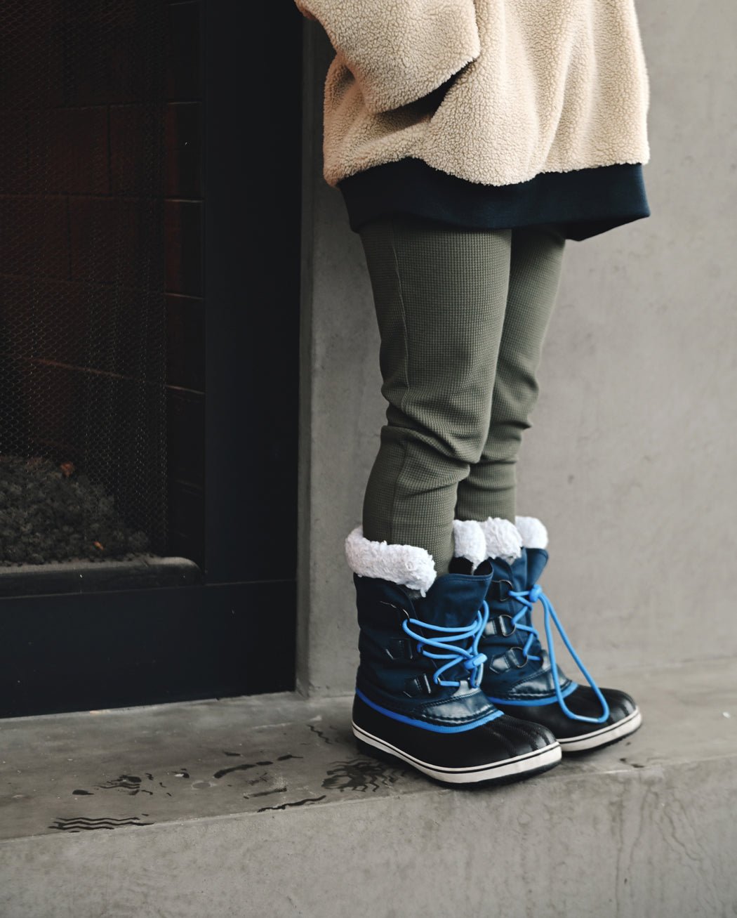 A child in Cozy Earth's Children's Bamboo Waffle Knit Base Layer Pant stands indoors near a fireplace, paired with a beige fleece jacket and blue winter boots with white fur trim on a concrete floor.