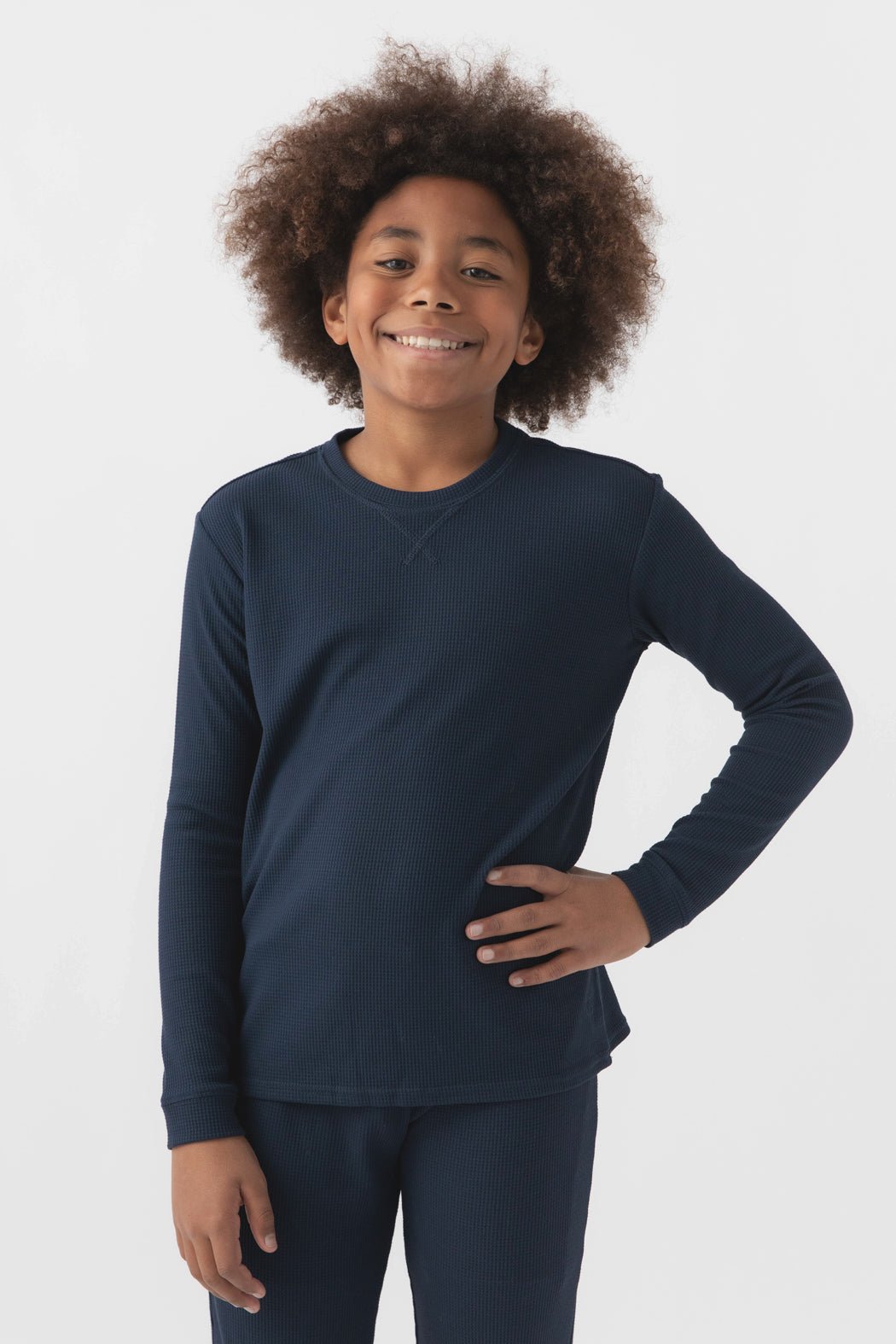 Smiling child with curly hair wears Cozy Earth's Children’s Bamboo Waffle Knit Base Layer Long Sleeve in navy blue, standing against a plain white background with one hand on their hip.|Color:Navy