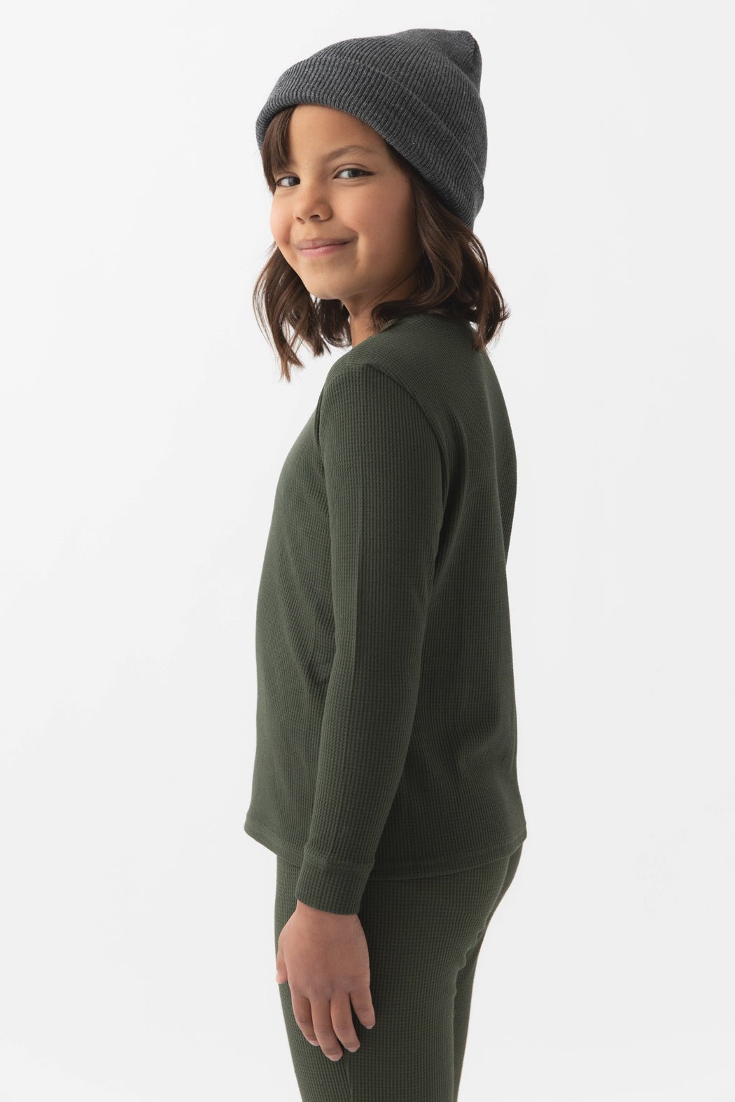 A young child stands sideways, smiling at the camera in a Cozy Earth Children's Bamboo Waffle Knit Base Layer Long Sleeve against a plain white background.