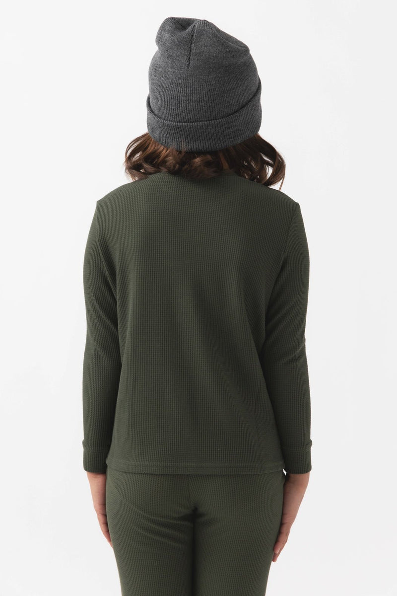 A child faces away from the camera, wearing Cozy Earth's Children's Bamboo Waffle Knit Base Layer Long Sleeve in dark green, with matching pants and a gray knit beanie against a plain white background.