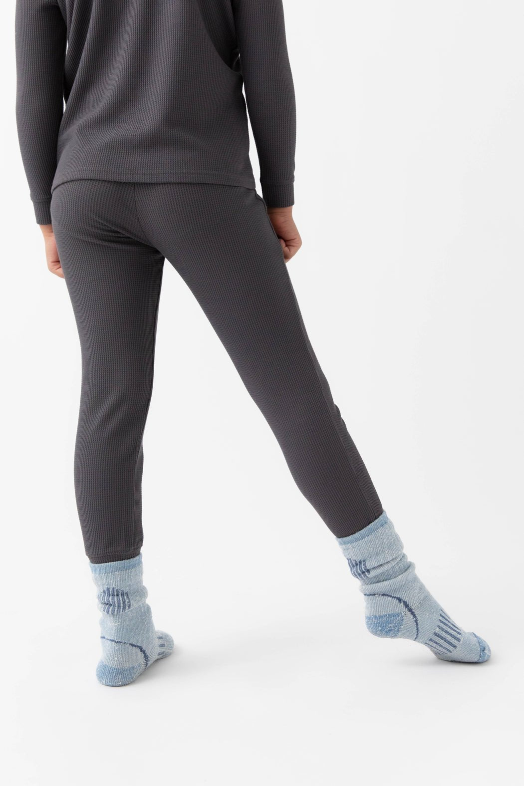 A child stands with their back turned, wearing Cozy Earth's Children's Bamboo Waffle Knit Base Layer Pant in dark ribbed fabric, paired with a matching top and light blue slouchy socks, set against a plain white background.