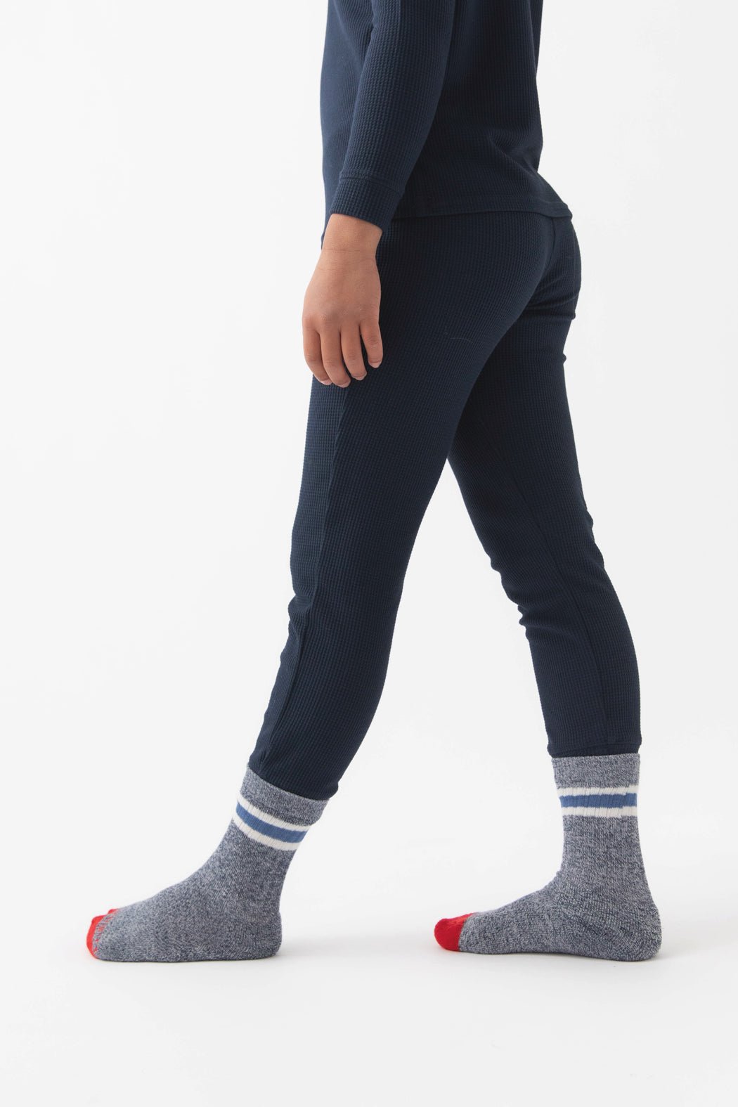 A child models the Children's Bamboo Waffle Knit Base Layer Pant by Cozy Earth, standing sideways in navy pajamas and gray socks with white stripes and red toes against a plain white background.