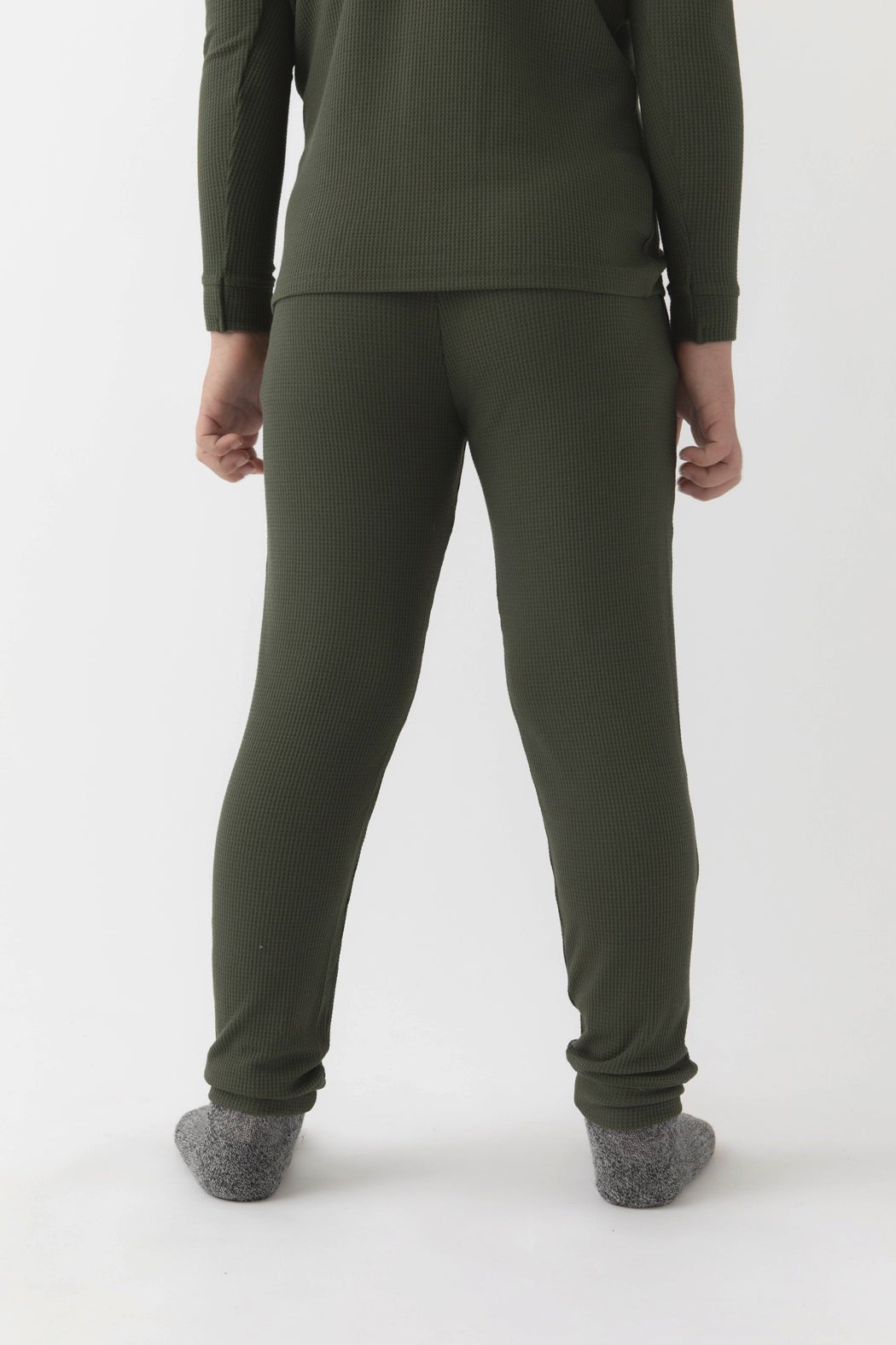 A child faces away from the camera wearing Cozy Earth's Children's Bamboo Waffle Knit Base Layer Pant in olive green, paired with a matching ribbed long-sleeve top and gray socks, set against a plain white background.
