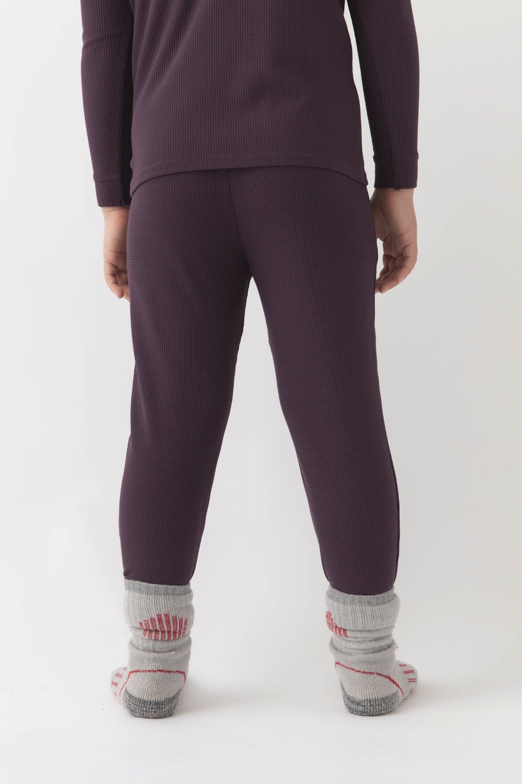 A child faces away, wearing Cozy Earth's dark purple Children's Bamboo Waffle Knit Base Layer Pant with a matching top and thick gray socks with red accents. The background is plain white.