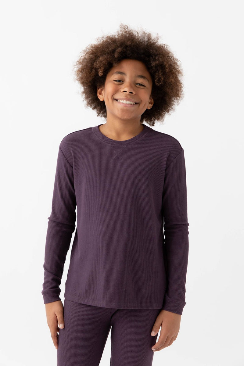 A smiling child with curly hair stands against a white background, wearing the Cozy Earth Children's Bamboo Waffle Knit Base Layer Long Sleeve in dark purple.