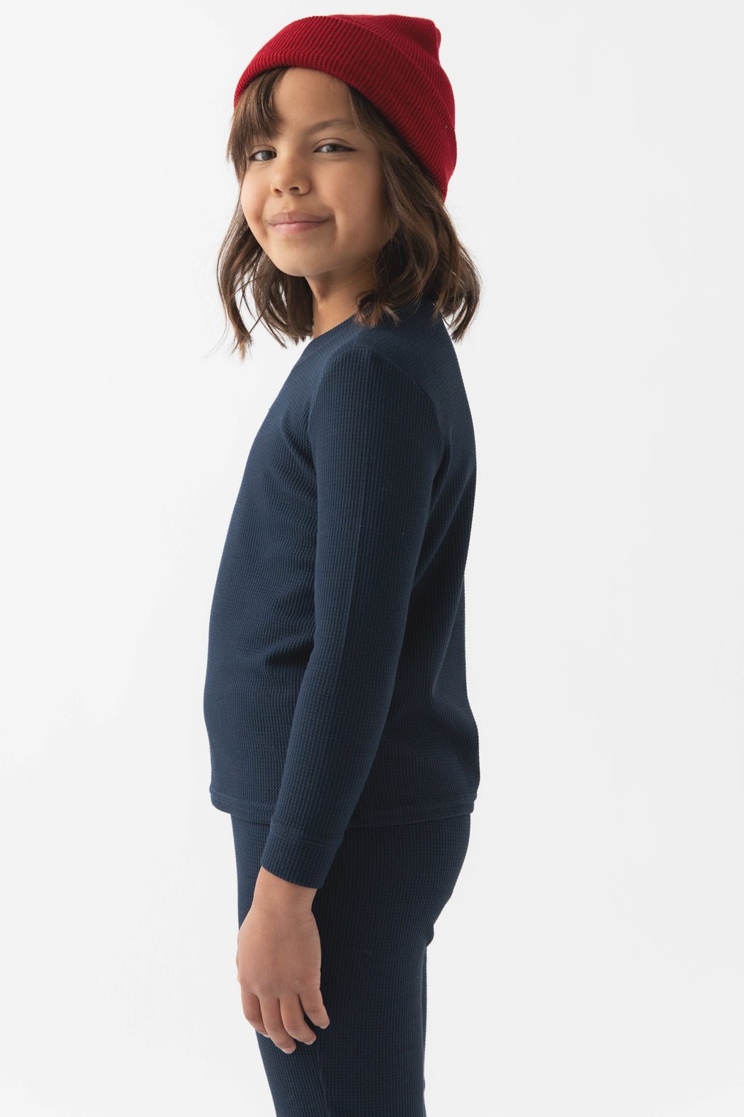 A young child with shoulder-length dark hair wears Cozy Earth's Children's Bamboo Waffle Knit Base Layer Long Sleeve in navy blue, paired with matching pants and a red knit beanie, standing sideways and smiling against a plain white background.