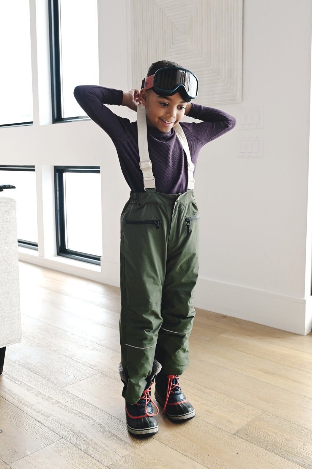 A child in green snow pants, snow boots, ski goggles, and a Cozy Earth Children's Bamboo Waffle Knit Base Layer Long Sleeve smiles indoors with hands behind their head, ready for winter fun.