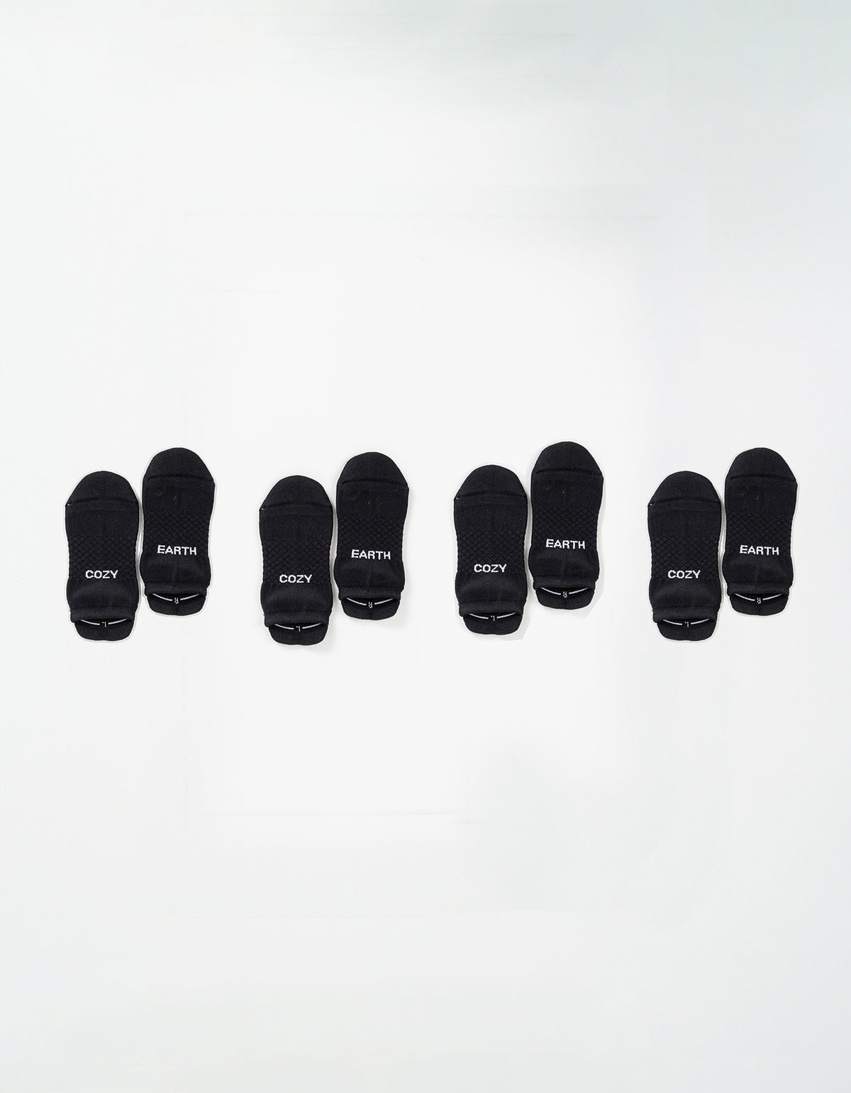 Four pairs of black Essential Ankle Socks by Cozy Earth are arranged in a row on a white background, each pair featuring "COZY" and "EARTH" in white text on the toes. 