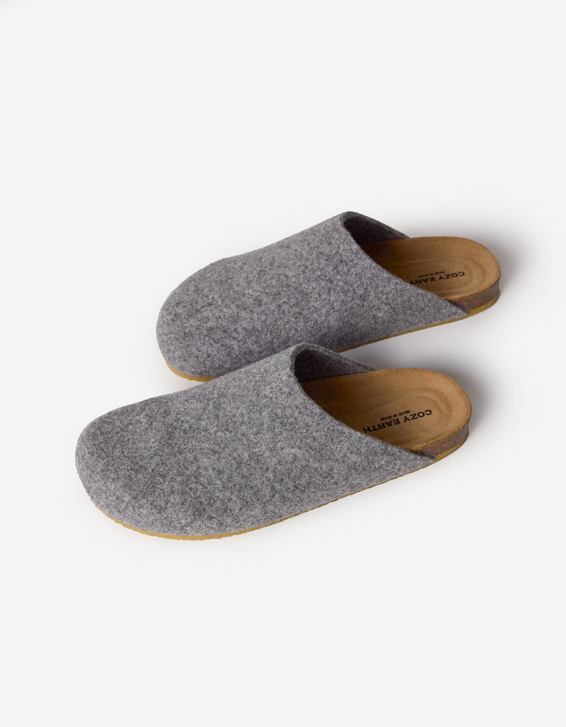 The Lakehouse Clog by Cozy Earth features a minimalist design with grey woolen uppers and natural brown soles. It has a closed toe, open back, and is ideal for indoor wear. A logo is visible on the insole, all showcased on a white background.