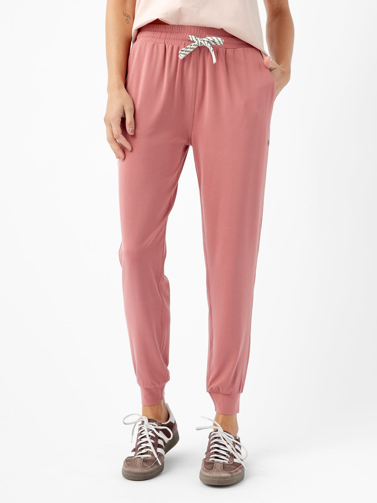 A person is standing and wearing Cozy Earth's Women's Studio Jogger, which features a pink color with a white drawstring and pockets where their hands rest. They are also dressed in a light-colored top and grey sneakers with white laces against a white background. 