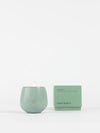 A pale green Cozy Earth Candle UNWIND in a round container, with a small flame, sits beside its matching box labeled "UNWIND" and "Cozy Earth" on a plain white background.