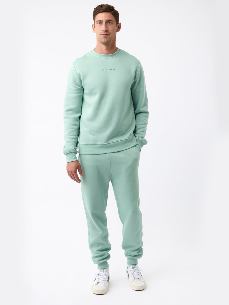 A man is standing against a plain white background, wearing the Men's CityScape Jogger set from Cozy Earth in light green, paired with white sneakers. His hands are relaxed by his sides, and he has short brown hair.