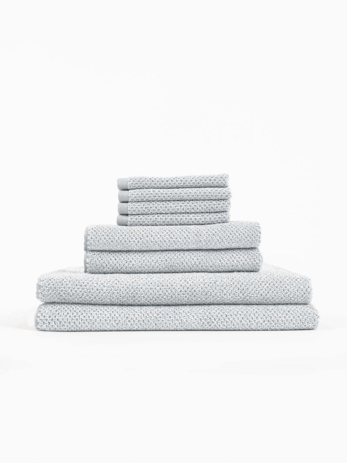 The Cozy Earth Nantucket Bath Towel Set features light grey textured towels in multiple sizes, neatly stacked from largest to smallest against a plain white background.|Color:Heathered Harbor Mist