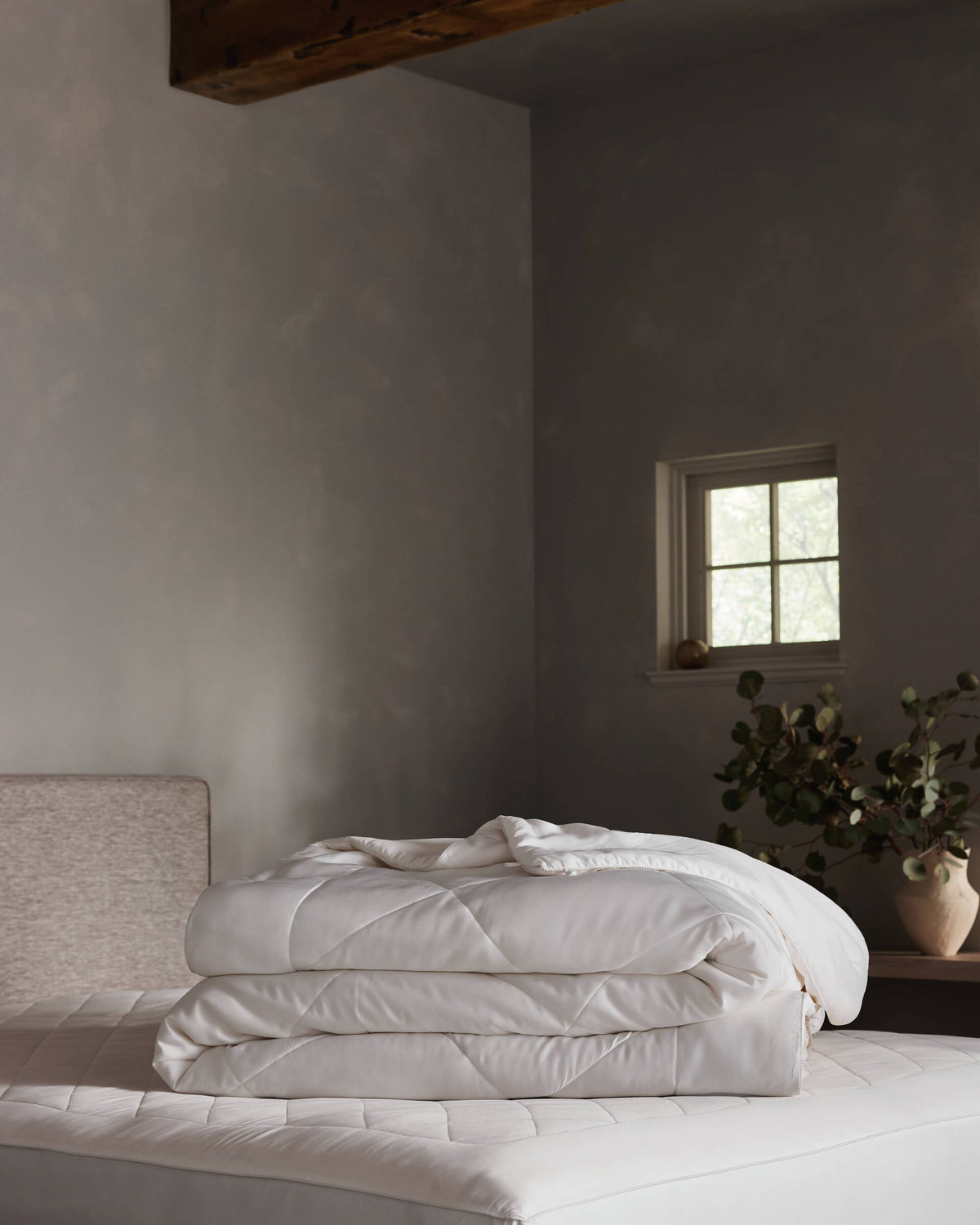 The Cozy Earth Bamboo Insert Bundle is neatly folded on a bed in a minimal, softly lit bedroom with gray walls, a small window, and a vase of greenery on the bedside table.|Filling:Standard