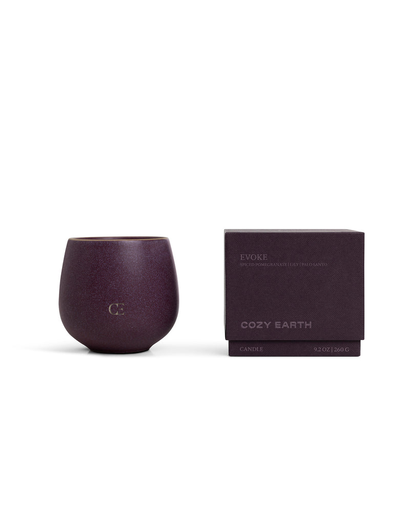 A deep purple Cozy Earth Candle EVOKE in a rounded cup with "CE" embossed on the front sits beside a matching purple box labeled "Evoke." Size: 9.2 oz (260 g).