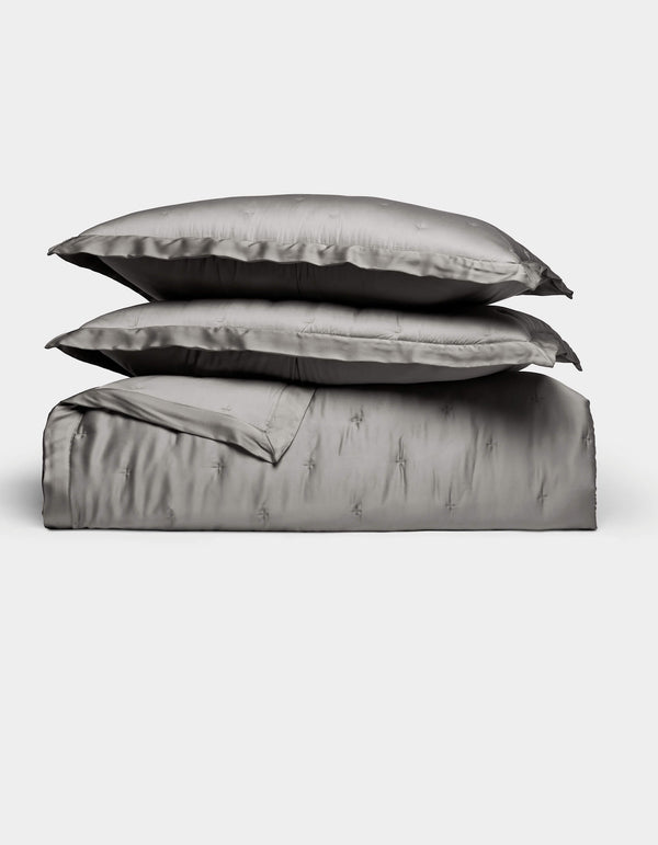 The Bamboo Quilt Bundle by Cozy Earth features lustrous silver-gray bed linens, including two stacked pillows, a folded sheet, and a duvet cover, all arranged against a plain white background.|Color:Charcoal