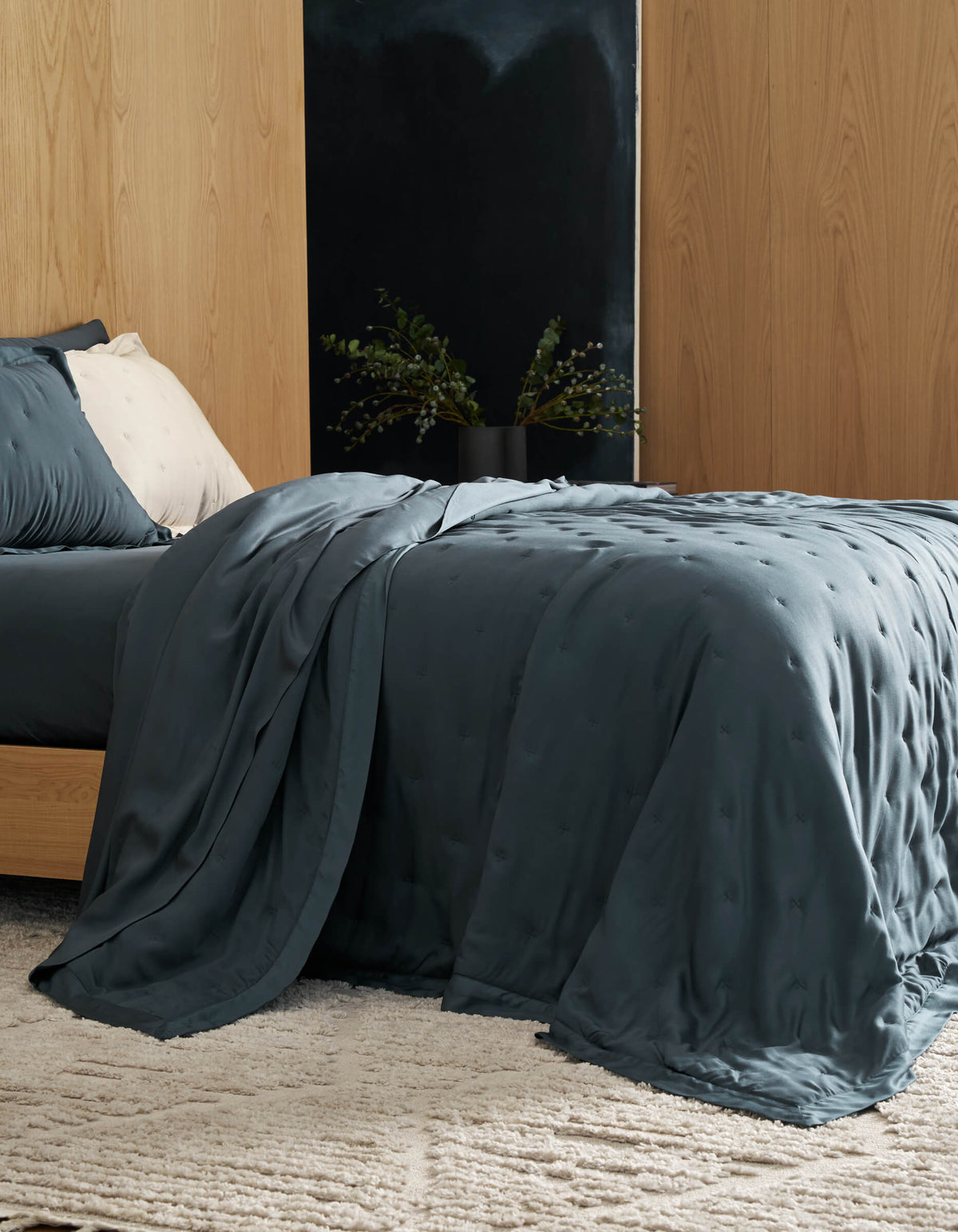 A modern bedroom features the Cozy Earth Bamboo Quilt Bundle on the bed, its dark blue texture matching the pillow. Light wood walls, a beige rug, and a black vase with green foliage contrast with the room’s neutral decor.