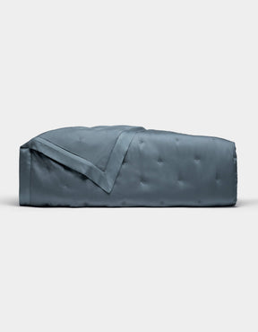 The Cozy Earth Bamboo Quilt, shown folded against a plain white background, features a blue-gray color with a subtle quilted pattern and smooth, shiny finish.|Color:Pacific Blue