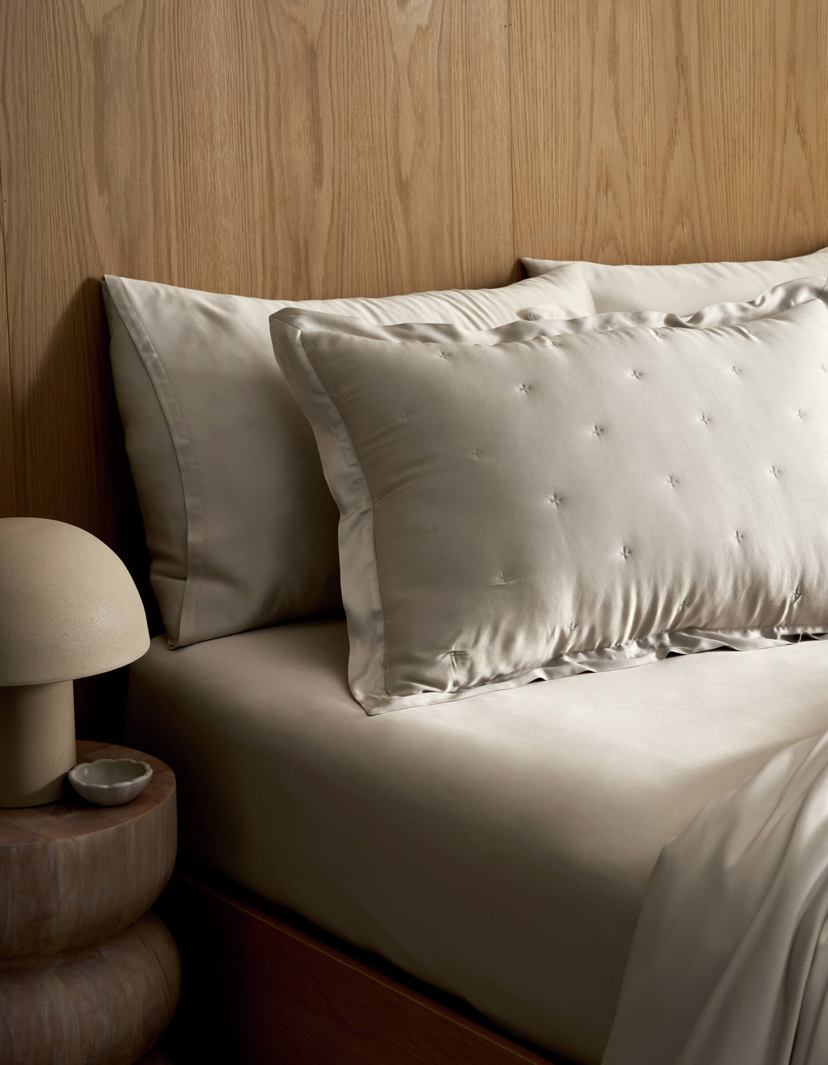 A neatly made bed features Cozy Earth's Bamboo Quilted Shams in light tones, set against a wooden headboard. A round bedside table holds a dome-shaped lamp and a small dish.