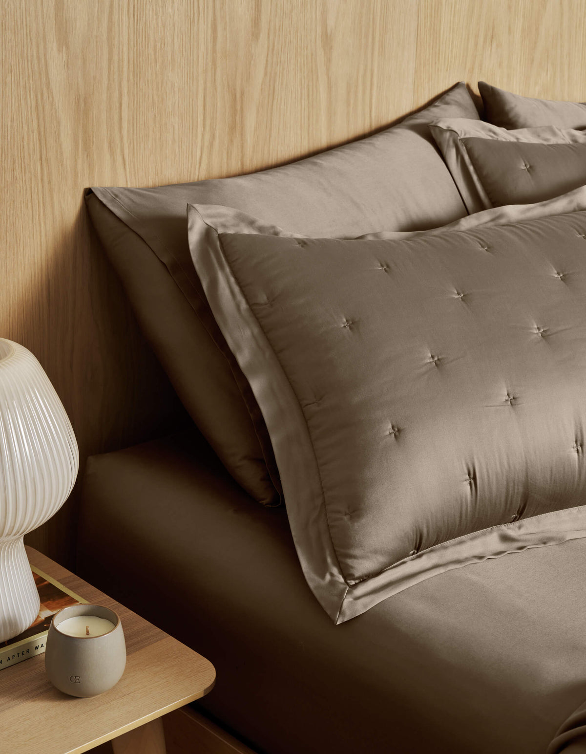 A neatly made bed with Cozy Earth Bamboo Quilted Shams and taupe satin bedding sits by a wooden nightstand topped with a ribbed white lamp, a lit candle, and a book against a light wood-paneled wall.