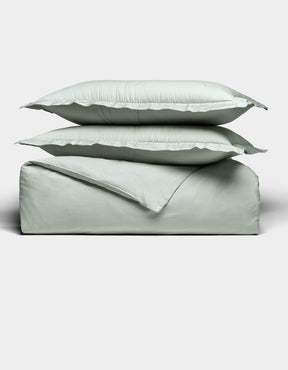 Two light gray pillows are stacked atop a matching folded Soft-Wash Cotton Duvet Set by Cozy Earth, all set against a plain white background. The bedding appears soft and smooth, highlighting its cozy and minimal aesthetic.|Color:Laurel