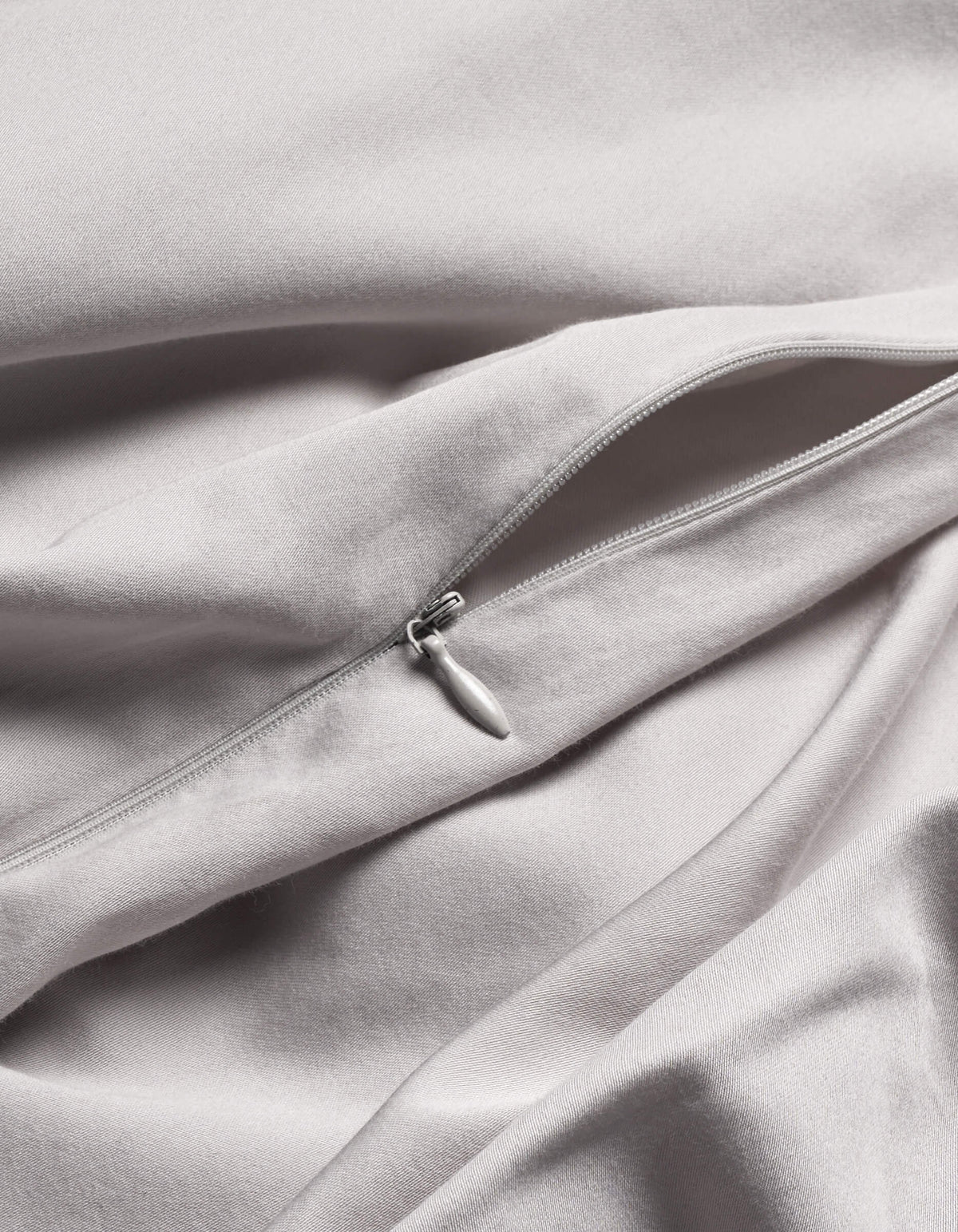 A close-up of Cozy Earth's Soft-Wash Cotton Duvet Cover in light grey shows a silver zipper partially unzipped on its smooth, gently folded fabric.