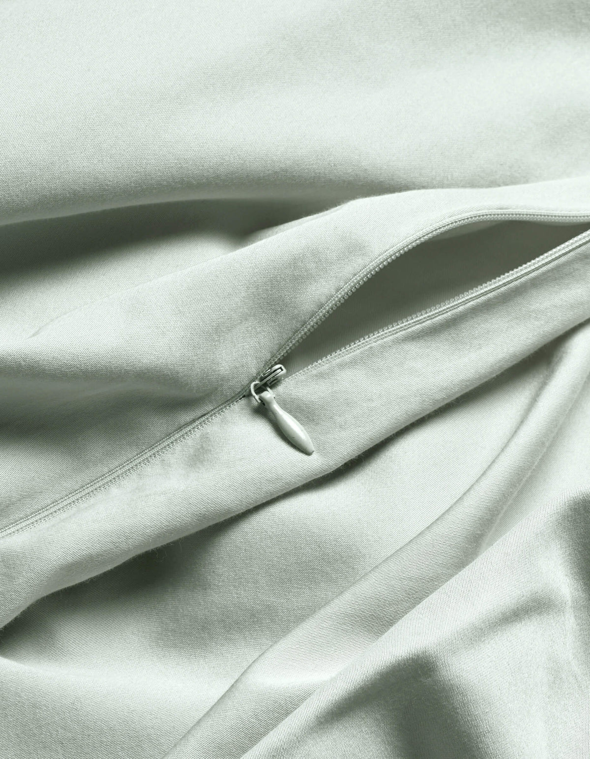 A close-up of the Cozy Earth Soft-Wash Cotton Duvet Cover in light gray or pale green shows a visible, partially unzipped zipper creating gentle folds and soft shadows on the smooth fabric.